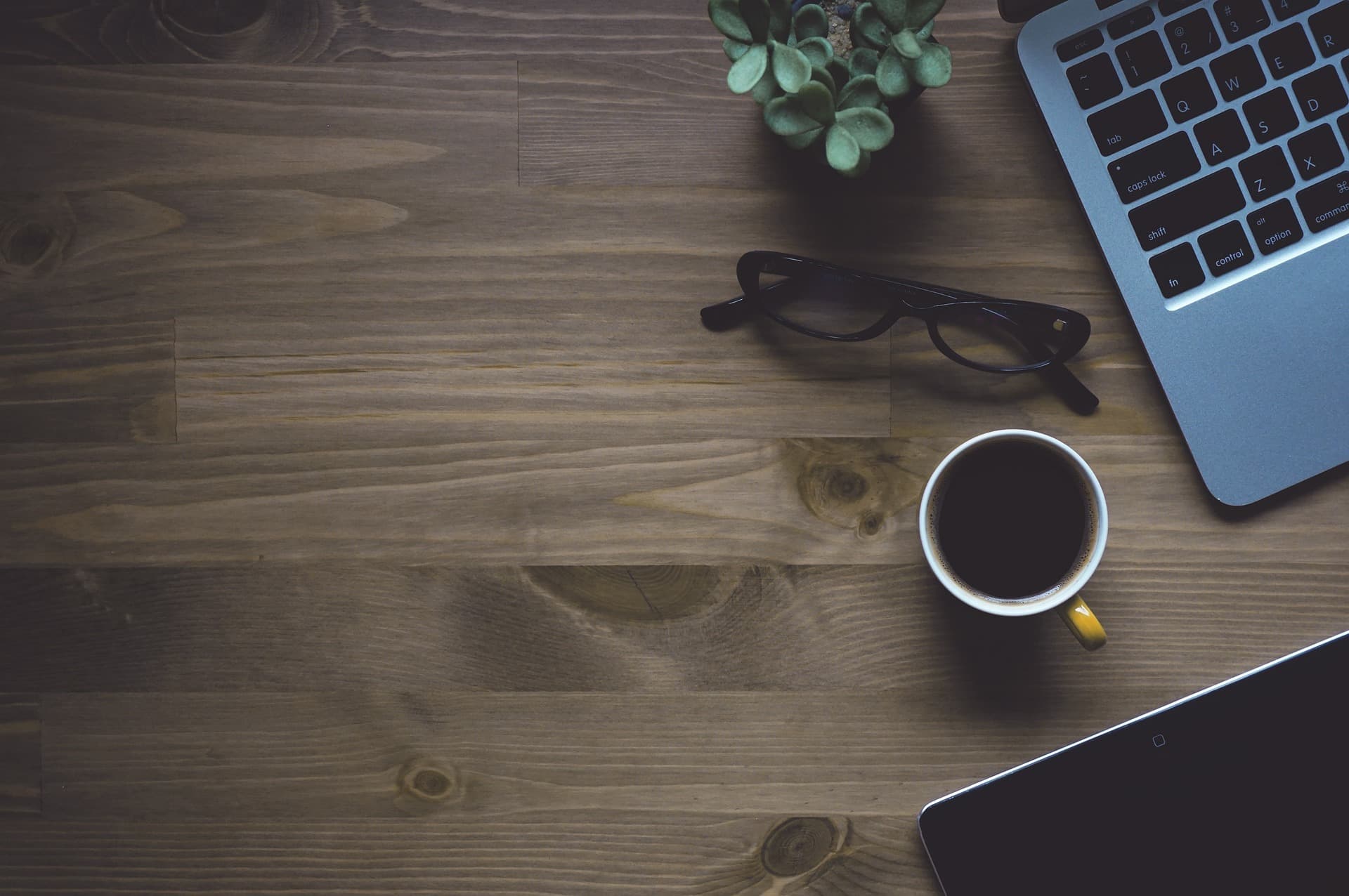 A wooden desk from above with a laptop, coffee, glasses, and a small plant